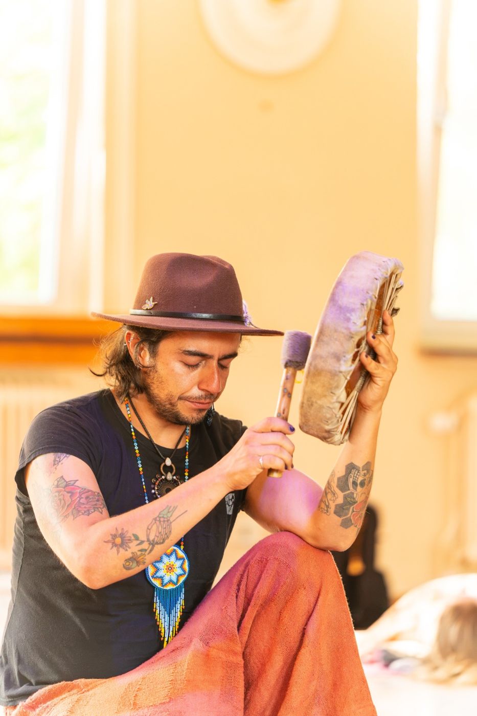 Ro in a brown hat, eyes closed, playing a frame drum